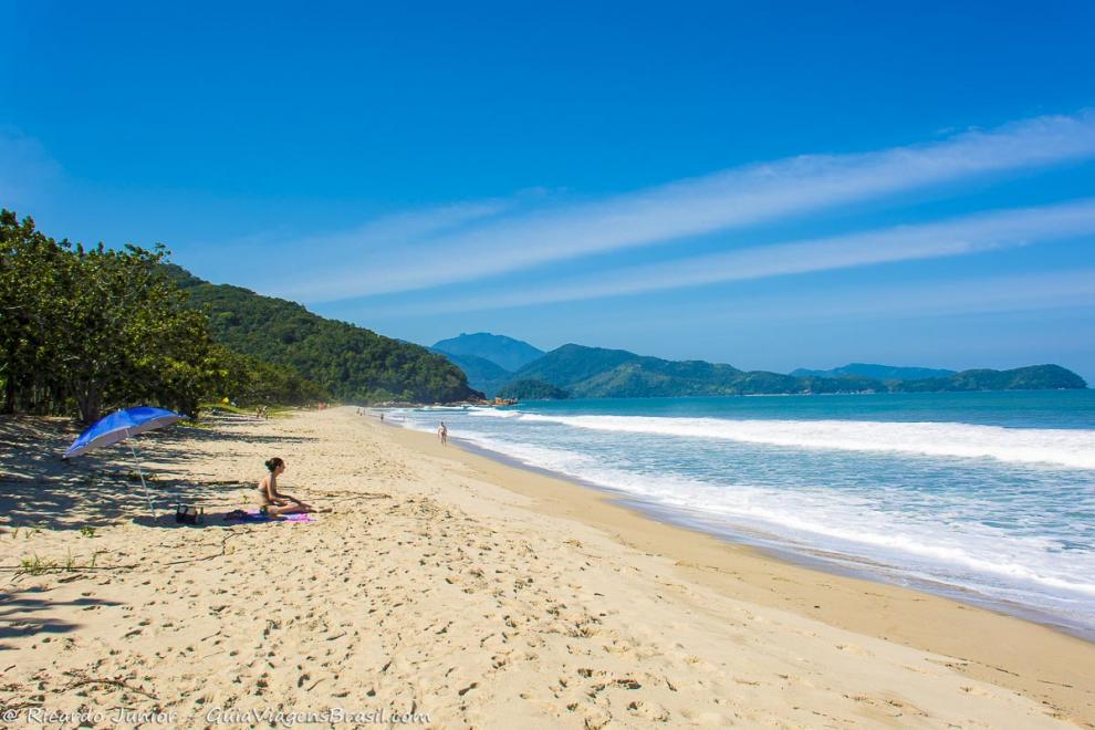 Imagem de uma moça sentada nas areias da praia olhando para o mar.